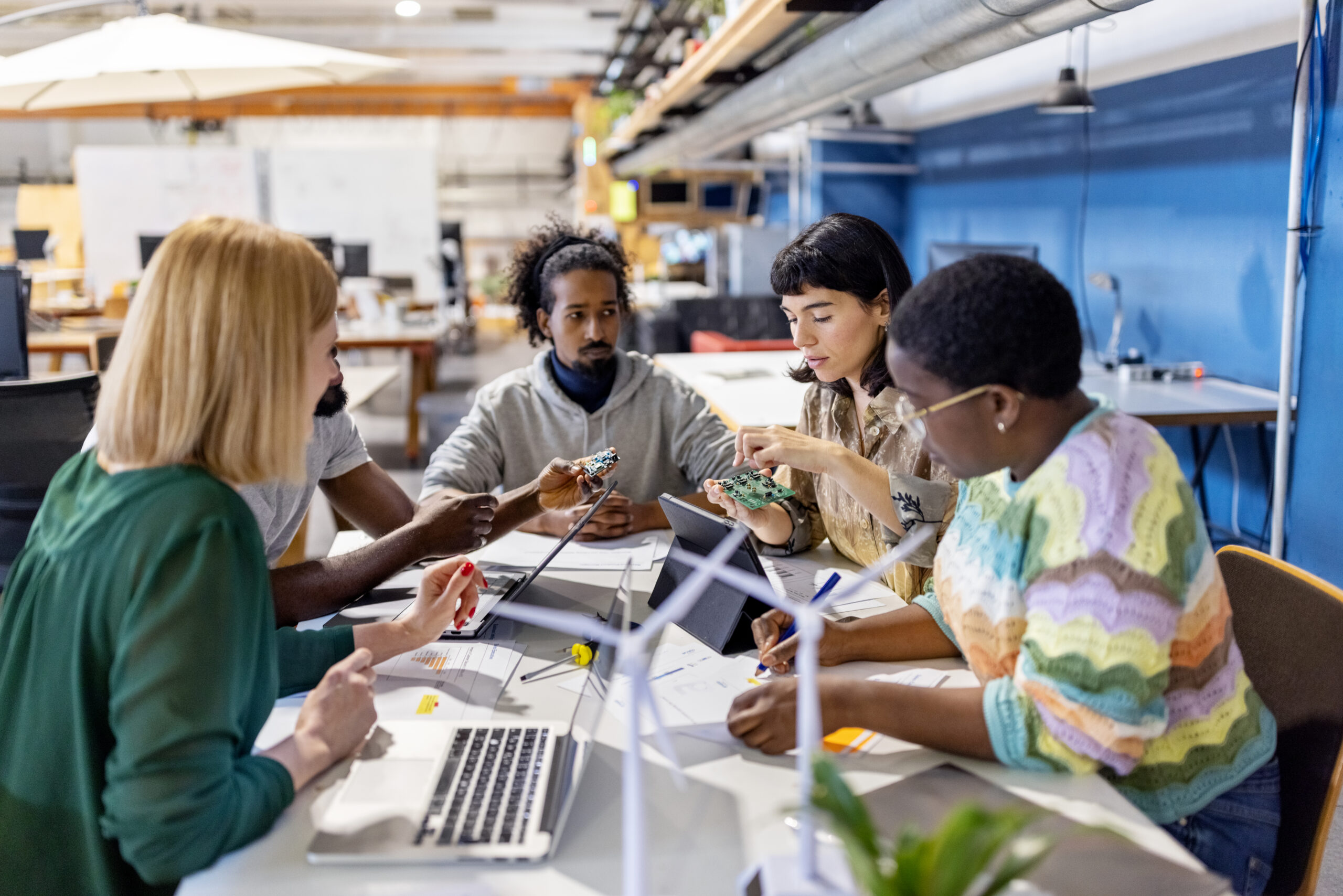 Young woman holding a PCB and discussing with colleagues at meeting at startup office. Group of young electronic engineers discussing designing a new circuit board for their project while sitting around a table at creative workspace.